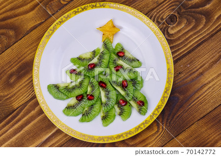 Christmas tree made of kiwi slices and pomegranate on wooden table. Top view. Creative idea for Christmas and New Year festive desserts. Funny food idea for kids Christmas tree made of kiwi slices and pomegranate on wooden table. Top view. Creative idea for Christmas and New Year festive desserts. Funny food idea for kids 70142772