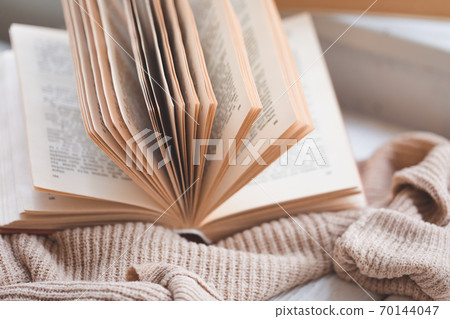 Warm and cosy hygge concept with white and beige sweater, opened book on a windowsill Warm and cosy hygge concept with white and beige sweater, opened book on a windowsill 70144047