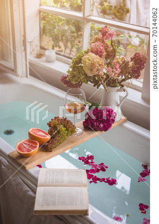 Bath tub with a tray with grapefruit slices, bunch of grapes, a glass of wine and a book, selective focus 70144062