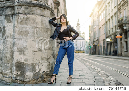 Full length portrait of young brunette Asian woman in jeans and stylish black leather jacket, sexy posing near old stone wall of the building, outdoor summer shot 70145736