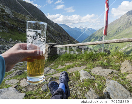 hiker holding beer glass with foot in rubber clogs relaxing at view from Nurnberger Hutte hut, valley with mountain peaks at Stubai hiking trail, Summer, Tyrol, Stubai Alps, Austria 70158889