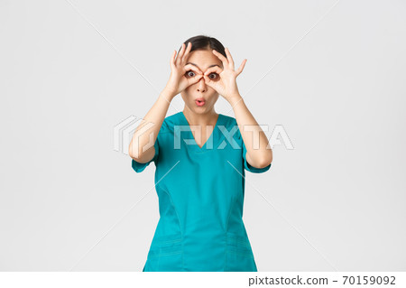 Covid-19, healthcare workers and preventing virus concept. Impressed funny and cute female asian nurse in scrubs looking through fingers over eyes as if stare in binoculats amazed 70159092