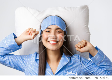 Close-up of happy and pleased smiling asian girl in blue pajamas and sleeping mask lying in bed on pillow, stretching satisfied and grinning after having good night sleep, waking up energized 70159160