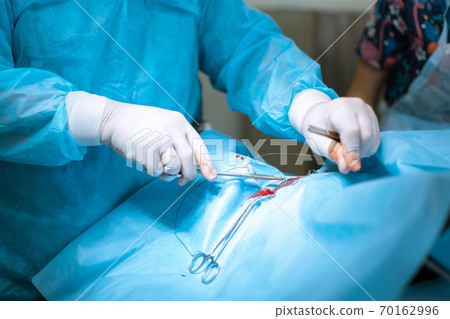 A male surgeon in a mask operates in a operating room with an assistant and an anesthesiologist. The medical team performs the operation. Gloved hands holding an insignificant plan. Tinted in blue 70162996