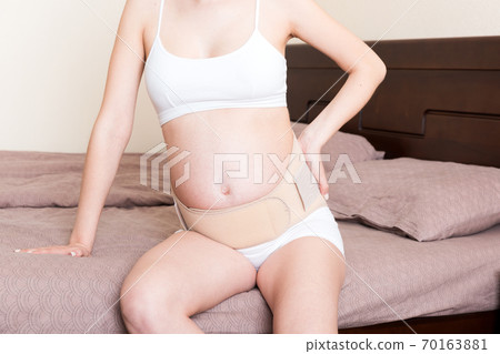 closeup pregnant woman dresses bandage on belly at home on bed. Orthopedic abdominal support belt closeup pregnant woman dresses bandage on belly at home on bed. Orthopedic abdominal support belt 70163881