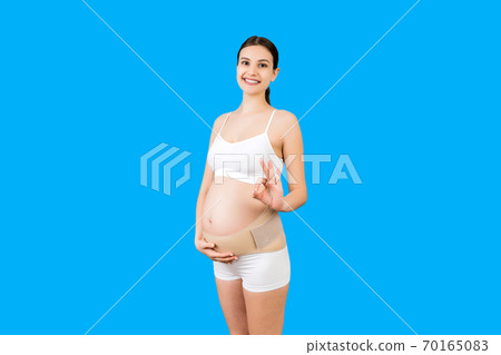 Portrait of pregnant woman in underwear on the third trimester wearing pregnancy belt and showing okay sign at blue background. Orthopedic abdominal support belt concept with copy space Portrait of pregnant woman in underwear on the third trimester wearing pregnancy belt and showing okay sign at blue background. Orthopedic abdominal support belt concept with copy space 70165083