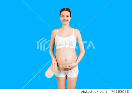 Portrait of pregnant woman in white underwear dressing maternity belt at blue background with copy space. Support belt against backache. Orthopedic abdominal support belt concept Portrait of pregnant woman in white underwear dressing maternity belt at blue background with copy space. Support belt against backache. Orthopedic abdominal support belt concept 70165088