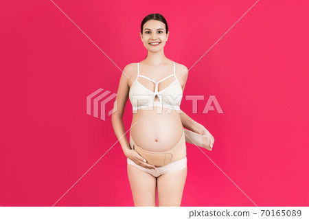 Portrait of pregnant woman in underwear dressing elastic maternity band at pink background with copy space. Orthopedic abdominal support belt concept Portrait of pregnant woman in underwear dressing elastic maternity band at pink background with copy space. Orthopedic abdominal support belt concept 70165089