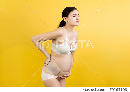 Portrait of pregnancy belt dressed on suffering pregnant woman in underwear for reducing pain in the back at yellow background with copy space. Orthopedic abdominal support belt concept Portrait of pregnancy belt dressed on suffering pregnant woman in underwear for reducing pain in the back at yellow background with copy space. Orthopedic abdominal support belt concept 70165108