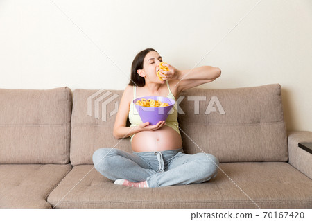 Pregnant woman sitting on the sofa enjoys eating chips from a bowl at home. Feeling hungry during pregnancy concept Pregnant woman sitting on the sofa enjoys eating chips from a bowl at home. Feeling hungry during pregnancy concept 70167420