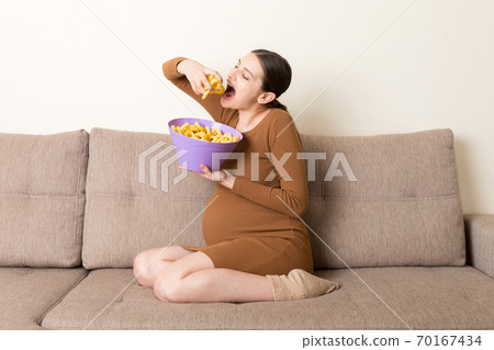 Pregnant woman sitting on the sofa enjoys eating chips from a bowl at home. Feeling hungry during pregnancy concept 70167434