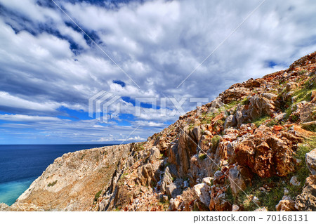 Beautiful wild red cliffs against clear blue sky and dynamic clouds. Diagonal compositition. Typical Greek or Cretan landscape. Mediterranean islands. Beautiful wild red cliffs against clear blue sky and dynamic clouds. Diagonal compositition. Typical Greek or Cretan landscape. Mediterranean islands. 70168131