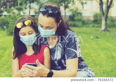Mom with daughter in the park in medical masks. Family of mom and kid wearing protective medical mask for prevent virus outdoors in the park. new normal concept. copy space Mom with daughter in the park in medical masks. Family of mom and kid wearing protective medical mask for prevent virus outdoors in the park. new normal concept. copy space 70168372