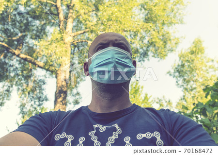 Portrait of young man wearing medical mask posing in sunny park. Close up portrait of a man looking away thoughtfully, wearing medical protective mask outdoors in the park. toned Portrait of young man wearing medical mask posing in sunny park. Close up portrait of a man looking away thoughtfully, wearing medical protective mask outdoors in the park. toned 70168472