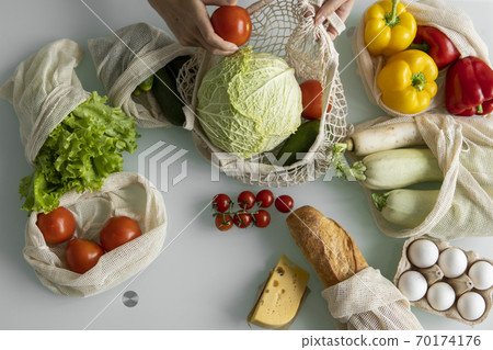 Woman came back from a market and unpacks a reusable grocery bag full of vegetables on a kitchen at home. Zero waste and plastic free concept. Girl is holding mesh cotton shopper with vegetables. Woman came back from a market and unpacks a reusable grocery bag full of vegetables on a kitchen at home. Zero waste and plastic free concept. Girl is holding mesh cotton shopper with vegetables. 70174176