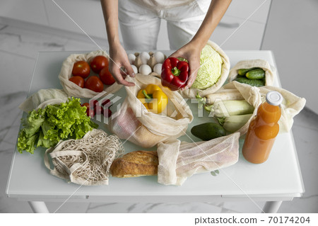 Woman's hand, holding a reusable grocery bag with vegetables on a kitchen at home and takes pepper out. Zero waste and plastic free concept. Mesh cotton shopper with vegetables. Ecology. Woman's hand, holding a reusable grocery bag with vegetables on a kitchen at home and takes pepper out. Zero waste and plastic free concept. Mesh cotton shopper with vegetables. Ecology. 70174204