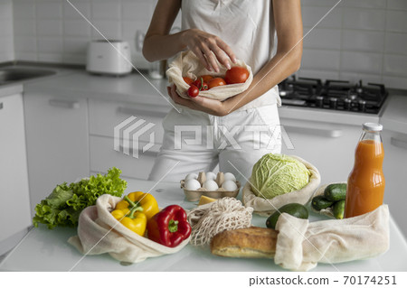 Woman's hand, holding a reusable grocery bag with vegetables on a kitchen at home and takes tomato out. Zero waste and plastic free concept. Mesh cotton shopper with vegetables. Ecology. Woman's hand, holding a reusable grocery bag with vegetables on a kitchen at home and takes tomato out. Zero waste and plastic free concept. Mesh cotton shopper with vegetables. Ecology. 70174251