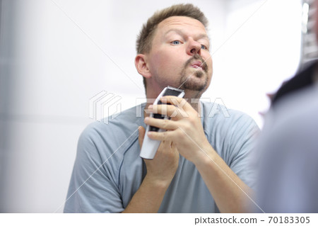 Young man shaves his face with razor while looking in mirror in bathroom 70183305