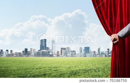 Green meadow behind red curtain and hand holding it Green meadow behind red curtain and hand holding it 70186750