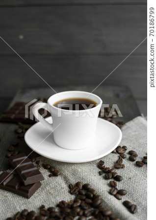 Coffee cup with cookies and chocolate with scattered coffee beans on linen and wooden table background. Mug of black coffee. Coffee cup with cookies and chocolate with scattered coffee beans on linen and wooden table background. Mug of black coffee. 70186799
