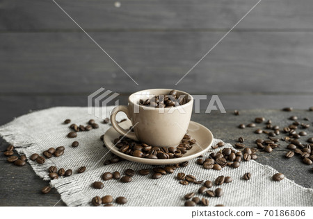 Coffee cup filled of fresh arabica or robusta coffee beans with scattered coffee beans on a linen textile and wood table. Coffee cup filled of fresh arabica or robusta coffee beans with scattered coffee beans on a linen textile and wood table. 70186806