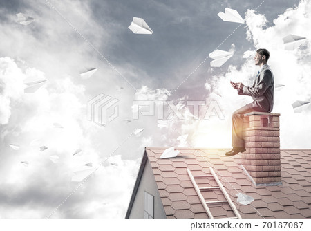 Man on brick roof reading book and paper planes flying in air Man on brick roof reading book and paper planes flying in air 70187087