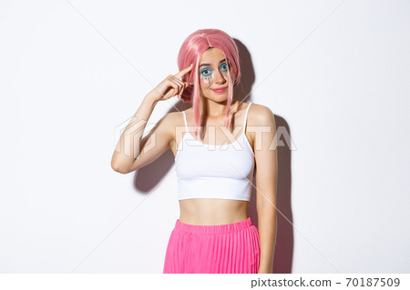 Portrait of sarcastic girl with pink hair and halloween makeup, scolding someone, pointing finger at head and looking at someone crazy or stupid, standing over white background 70187509