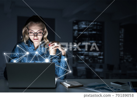 Attractive blonde wearing glasses in dark office using laptop. Mixed media Attractive blonde wearing glasses in dark office using laptop. Mixed media 70189058