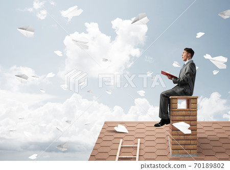 Man on brick roof reading book and paper planes flying in air Man on brick roof reading book and paper planes flying in air 70189802