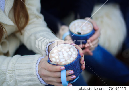 The hands of a girl and a guy are holding blue knitted mugs with cocoa and white marshmallows The hands of a girl and a guy are holding blue knitted mugs with cocoa and white marshmallows 70196625