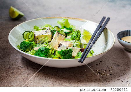 Green vegan salad with broccoli, smoked tofu and tahini dressing in white bowl, dark background. 70200411