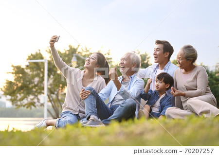 three generation asian family sitting on grass taking a selfie using mobile phone outdoors in park three generation asian family sitting on grass taking a selfie using mobile phone outdoors in park 70207257