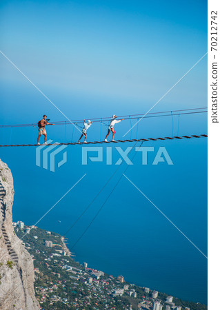 Family crossing the chasm on the rope bridge. Black sea background, Crimea, Russia Family crossing the chasm on the rope bridge. Black sea background, Crimea, Russia 70212742