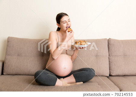 Pregnant woman enjoys eating cookies resting on the sofa at home. Unhealthy sweet pastry during pregnancy concept Pregnant woman enjoys eating cookies resting on the sofa at home. Unhealthy sweet pastry during pregnancy concept 70214553