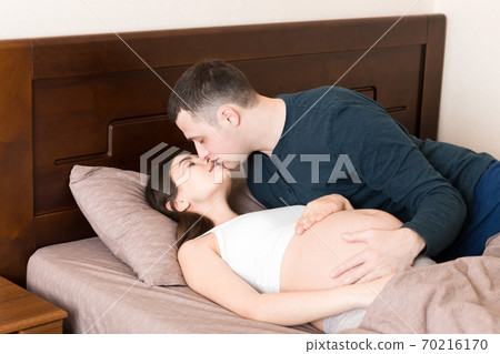 Young family. Attractive pregnant woman and man resting on bed and kissing. Happy pregnant family 70216170
