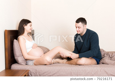 Happy pregnant family. Husband massaging his pregnant wife legs and sitting together on couch in their home 70216171