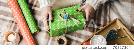 Young woman sits on plaid in cozy knitted woolen white sweater, socks and wraps Christmas gift in polka dot wrapping paper. Wooden tray with mug of cocoa with marshmallows, toy tree, candle, straws. Young woman sits on plaid in cozy knitted woolen white sweater, socks and wraps Christmas gift in polka dot wrapping paper. Wooden tray with mug of cocoa with marshmallows, toy tree, candle, straws. 70217304