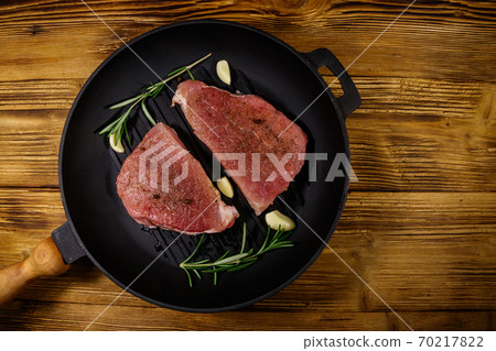 Raw pork steaks ready for preparation with rosemary, garlic and spices in cast iron grill frying pan on wooden table. Top view Raw pork steaks ready for preparation with rosemary, garlic and spices in cast iron grill frying pan on wooden table. Top view 70217822