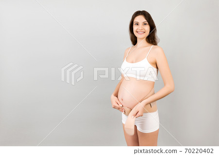 Portrait of pregnant woman in white underwear putting on a bandage at gray background with copy space. Orthopedic abdominal support belt concept Portrait of pregnant woman in white underwear putting on a bandage at gray background with copy space. Orthopedic abdominal support belt concept 70220402