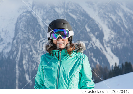 A woman in a colorful ski suit, helmet and sunglasses after skiing stands against the backdrop of mountain peaks. Sports concept, people, travel. 70220442