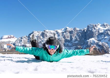 A teenage girl in a colorful ski suit, helmet and sunglasses lies in the snow and smiles happily against the backdrop of mountain peaks. Sports concept, people, travel. 70220452