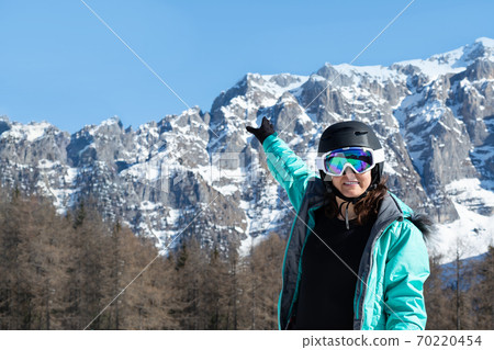A woman in a colorful ski suit, helmet and sunglasses after skiing stands against the backdrop of mountain peaks. Sports concept, people, travel. 70220454