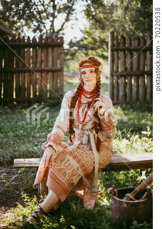 A beautiful Slavic girl with long blonde hair and brown eyes in a white and red embroidered suit is sitting next to a wooden fence.Traditional clothing of the Ukrainian region. A beautiful Slavic girl with long blonde hair and brown eyes in a white and red embroidered suit is sitting next to a wooden fence.Traditional clothing of the Ukrainian region. 70220538