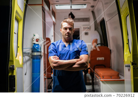 Man in a medical uniform looking at the camera seriously, arms folded, ambulance car in the background Man in a medical uniform looking at the camera seriously, arms folded, ambulance car in the background 70226912