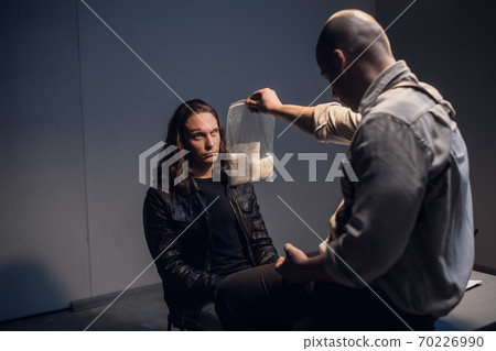 A detained white man with long hair receives irrefutable evidence of his guilt from a criminal investigator at the police station. A detained white man with long hair receives irrefutable evidence of his guilt from a criminal investigator at the police station. 70226990