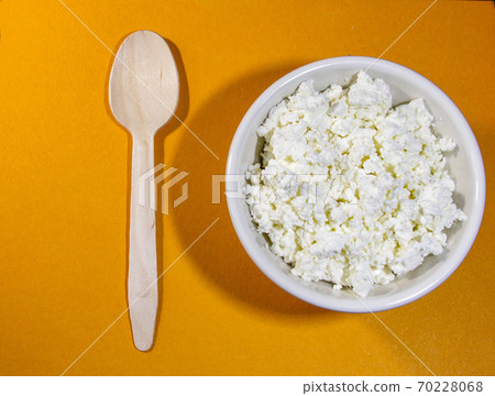 Cottage cheese in white bowl on orange table with spoon. Top view. 70228068