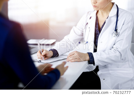 Unknown woman-doctor is writing some medical recommendations to her patient, while they are sitting together at the desk in the sunny cabinet in a clinic. Physician is using a clipboard, close-up Unknown woman-doctor is writing some medical recommendations to her patient, while they are sitting together at the desk in the sunny cabinet in a clinic. Physician is using a clipboard, close-up 70232777
