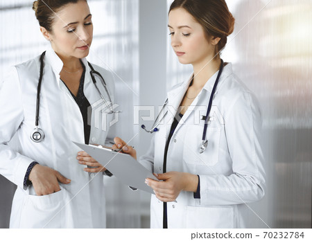 Two female physicians are discussing their patient's medical tests, while standing in a sunny clinic office. Doctors use a clipboard at work. Teamwork in medicine 70232784
