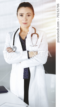 Smiling professional woman-doctor is standing with crossed arms in a sunny clinic office. Portrait of friendly female physician with a stethoscope at work. Perfect medical service in a hospital 70232786
