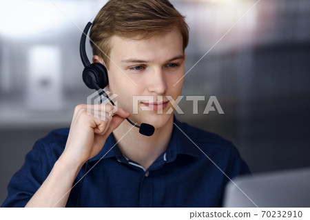 Young blond businessman using headset and computer in a darkened office, glare of light on the background. Startup business means working hard Young blond businessman using headset and computer in a darkened office, glare of light on the background. Startup business means working hard 70232910